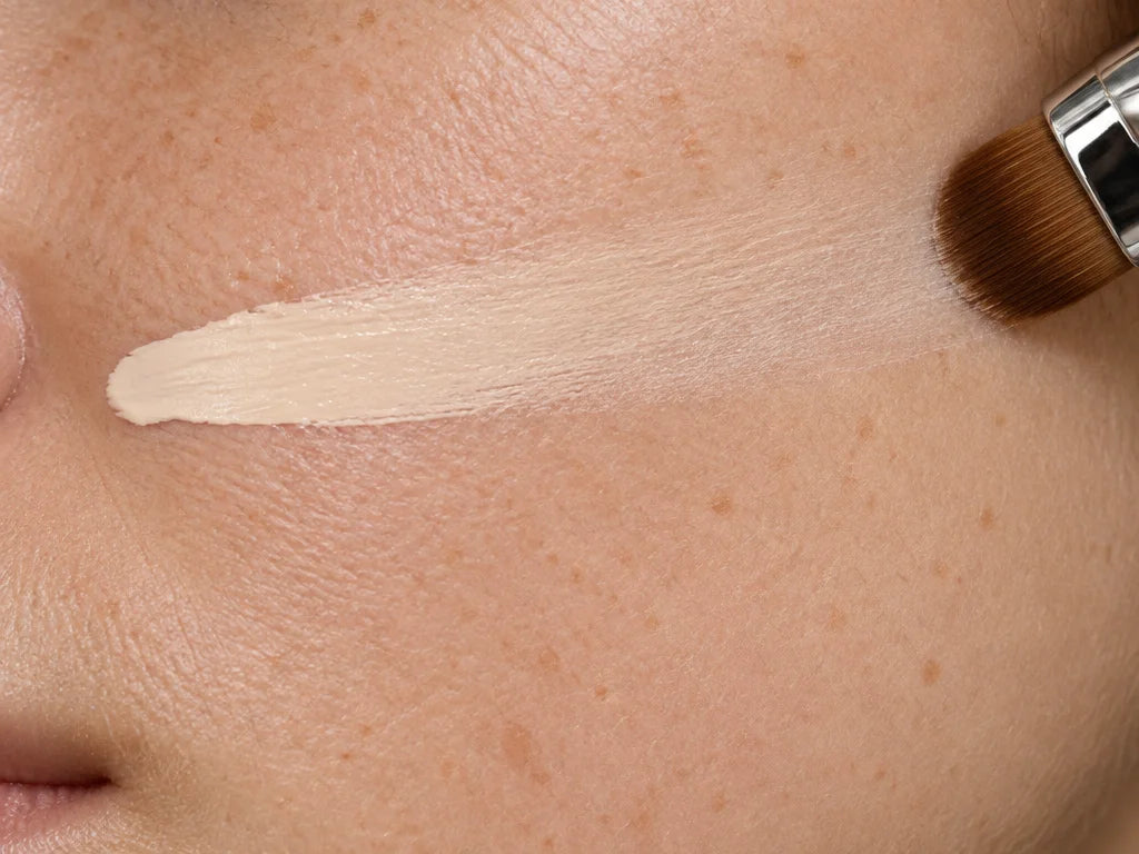 A horizontal close-up beauty photograph of a pale foundation-cream stroke on real cheek skin, the st